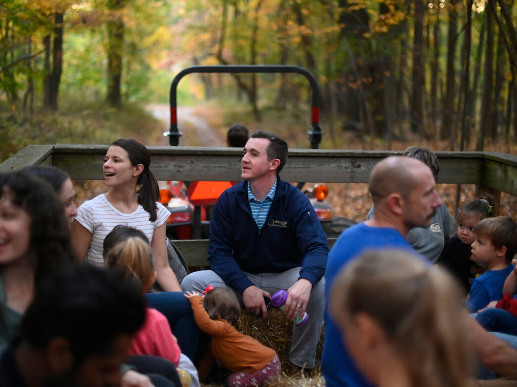 Families and children sit on hay bales in a tractor-pulled wagon during a fall hayride through a wooded trail at the Schuylkill Center.