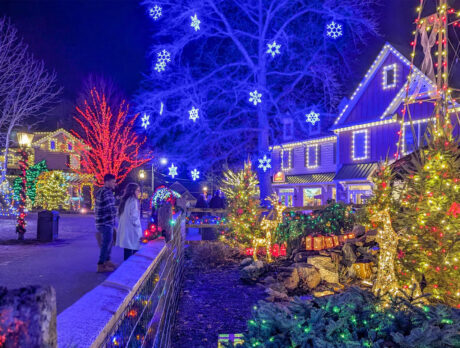 Two visitors admire the colorful holiday lights and glowing snowflake decorations illuminating the buildings and trees at Peddler's Village on a winter night.