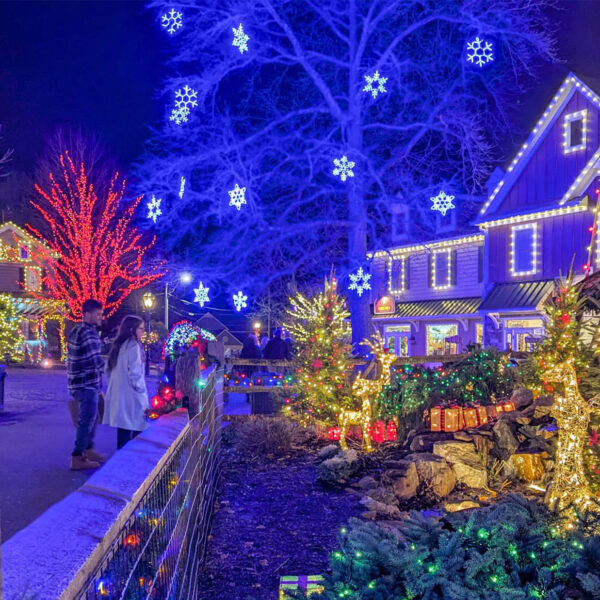 Two visitors admire the colorful holiday lights and glowing snowflake decorations illuminating the buildings and trees at Peddler's Village on a winter night.