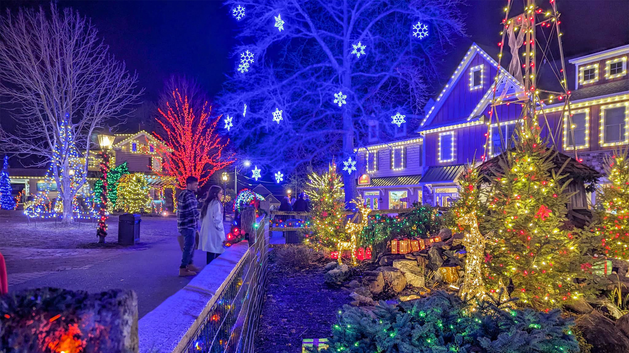 Two visitors admire the colorful holiday lights and glowing snowflake decorations illuminating the buildings and trees at Peddler's Village on a winter night.