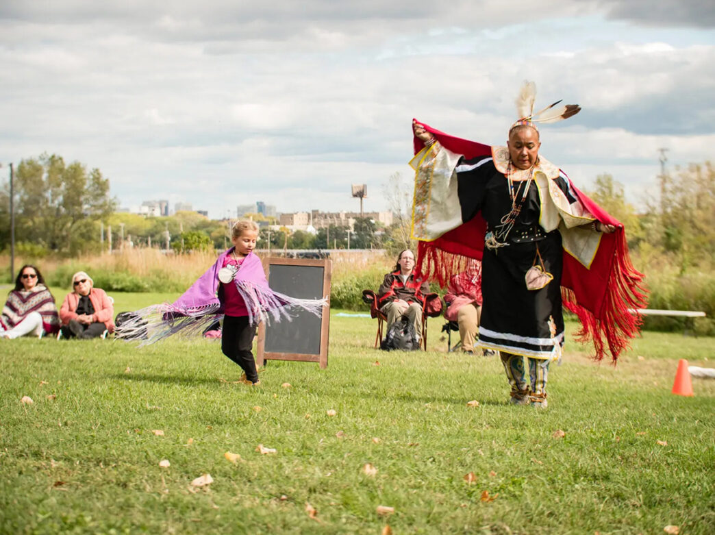 A dancer dressed in traditional Indigenous regalia performs alongside a child wrapped in purple shawl on a grassy lawn.