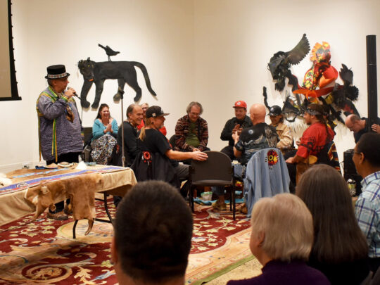 Attendees watch a Lenape cultural presentation featuring a drum circle and traditional regalia inside a gallery at Woodmere.