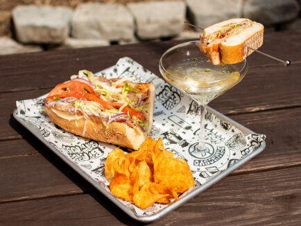A hoagie sandwich served with chips and a martini garnished with a mini hoagie on a picnic table at Liberty Kitchen.