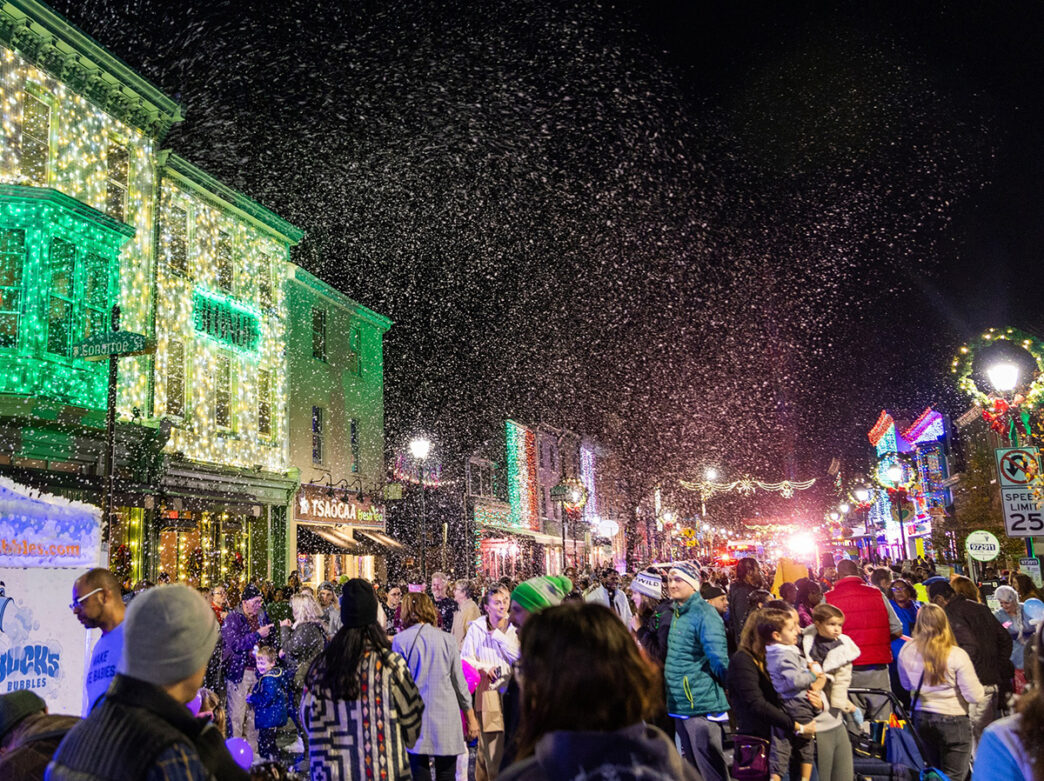 Crowds fill Main Street under a flurry of snow as colorful holiday lights illuminate the buildings.