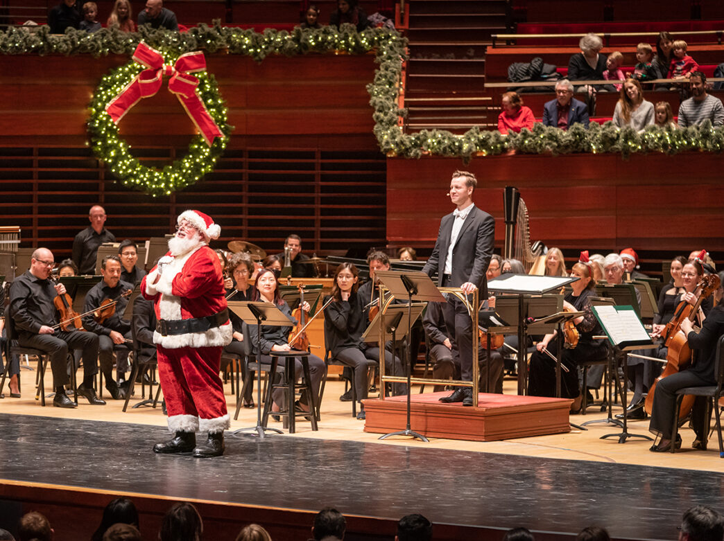 Santa Claus joins the Philadelphia Orchestra on stage during a festive holiday concert at the Kimmel Center, with a large illuminated wreath and garland in the background.