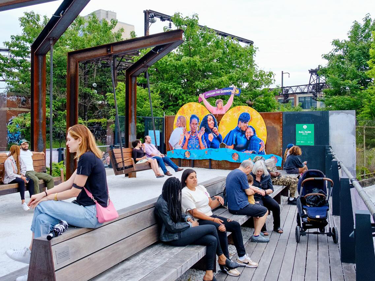 People relax on wooden benches and swings at The Rail Park, with trees and public art visible in the background.