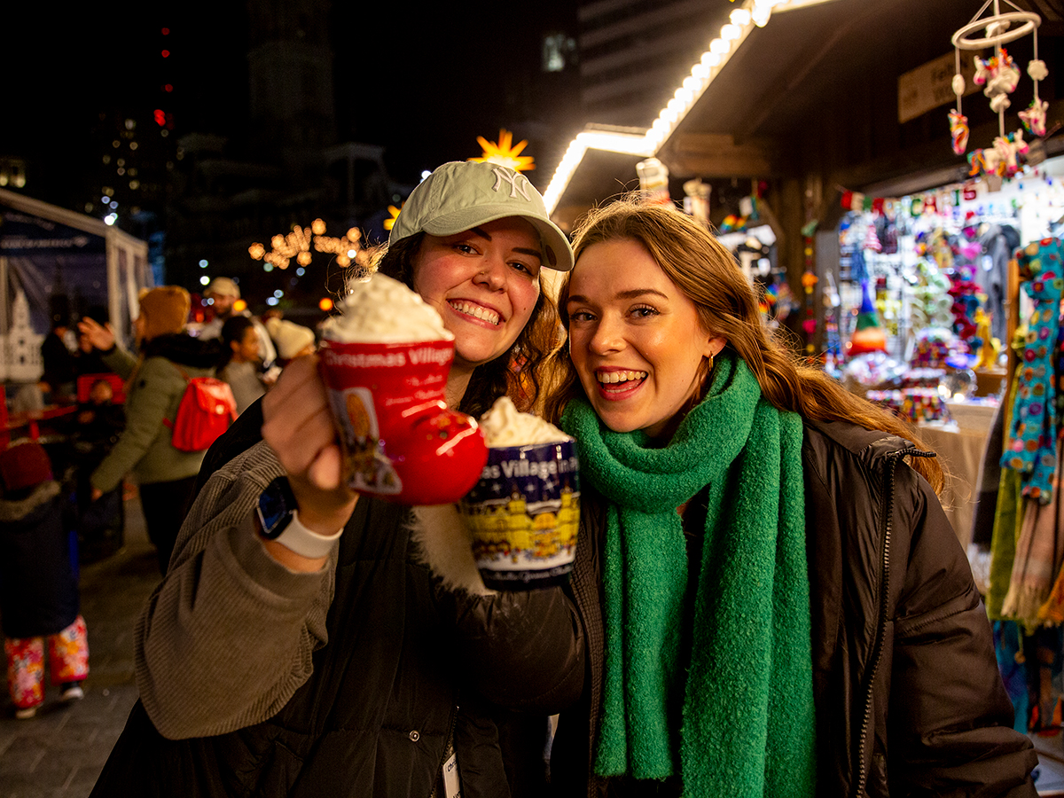 Two smiling women wearing winter jackets and scarfs hold up festive mugs topped with whipped cream at Christmas Village in Philadelphia.