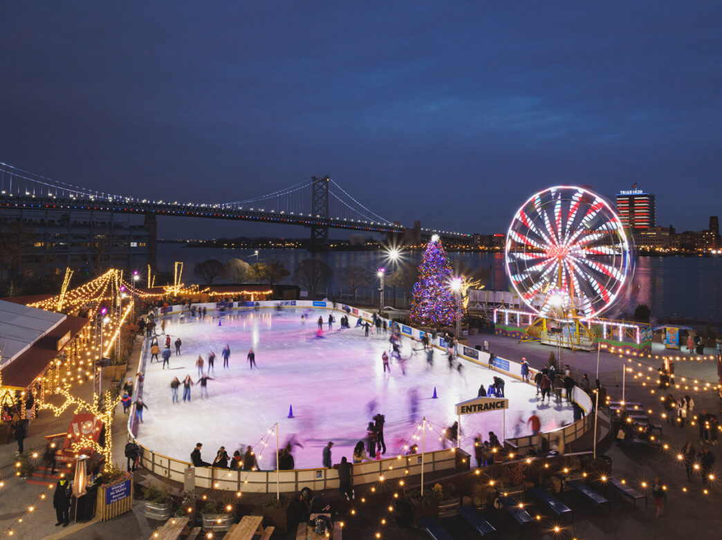 Skaters glide across an ice rink with a Ferris wheel and holiday tree sparkle against the Delaware River and Benjamin Franklin Bridge in the background.
