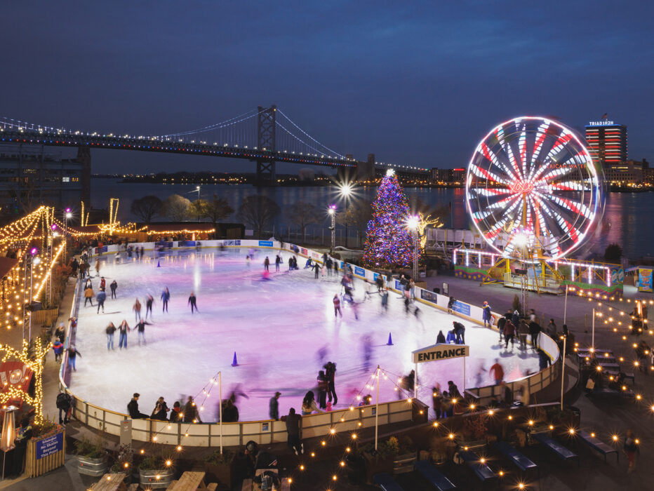Skaters glide across an ice rink with a Ferris wheel and holiday tree sparkle against the Delaware River and Benjamin Franklin Bridge in the background.