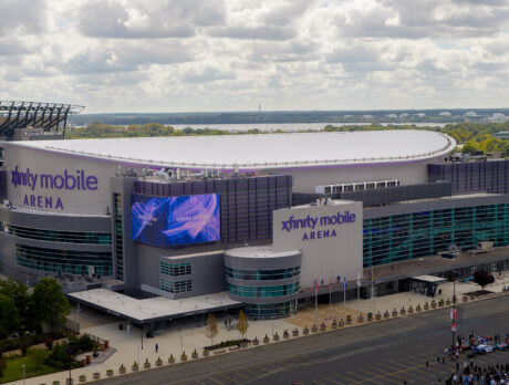 A drone photo of the sleek, modern Xfinity Mobile Arena with glass walls, flags flying out front and a bright purple marquee.