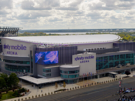 A drone photo of the sleek, modern Xfinity Mobile Arena with glass walls, flags flying out front and a bright purple marquee.