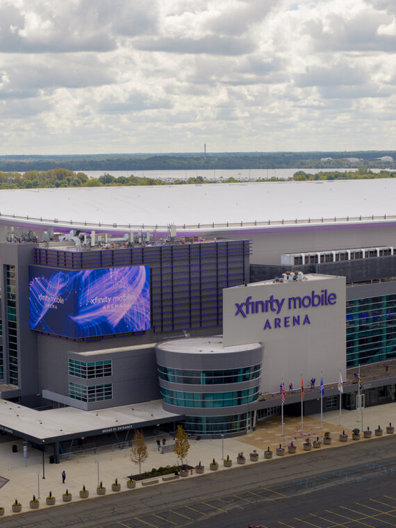 A drone photo of the sleek, modern Xfinity Mobile Arena with glass walls, flags flying out front and a bright purple marquee.