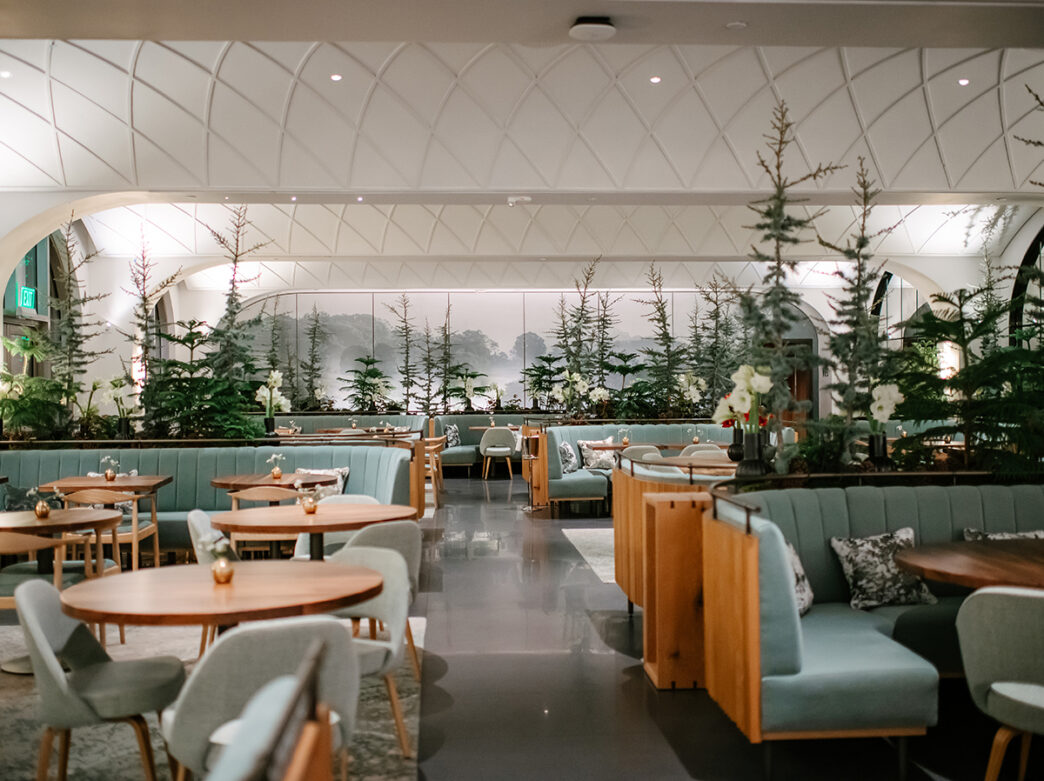 A spacious, elegant restaurant dining room with soft green banquettes, light wood tables and small evergreen trees arranged in the space under a vaulted white ceiling.