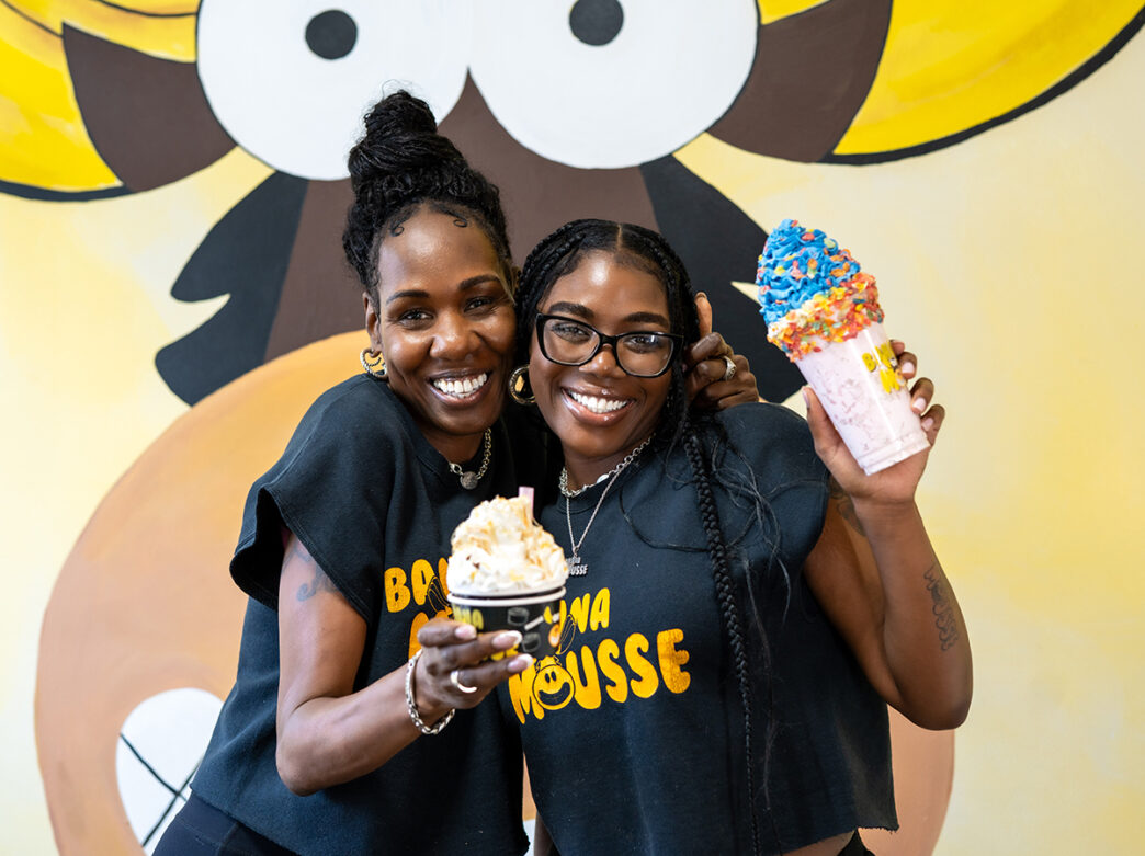 Two women in matching black Banana Mousse shirts smile and pose with a colorful milkshake and a cup of mousse in front of the shop's playful, cartoon style mural.