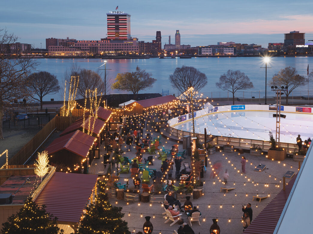 An evening aerial view of an ice rink, fire pits and string lights along the Delaware River waterfront.