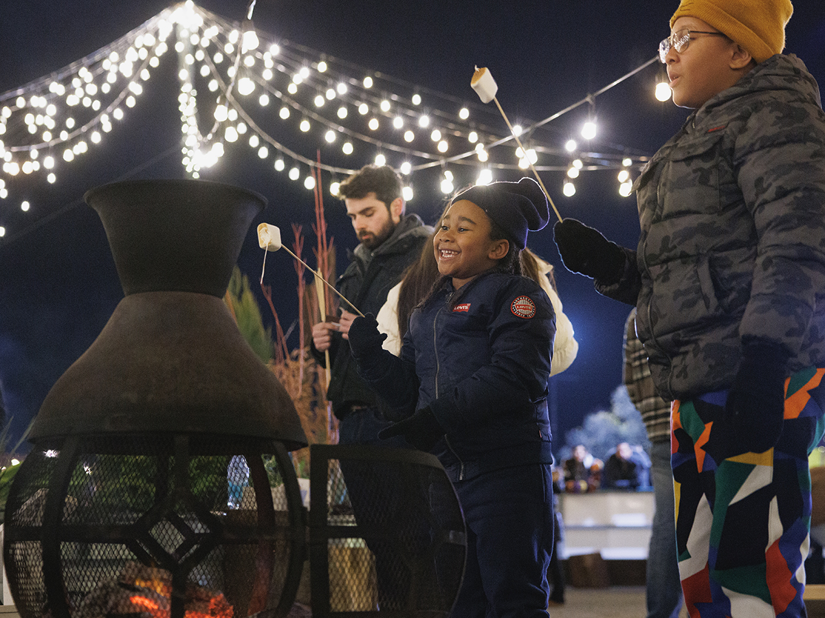 Children wearing winter jackets, hats and gloves roast marshmallows over a fire pit under a canopy of string lights.