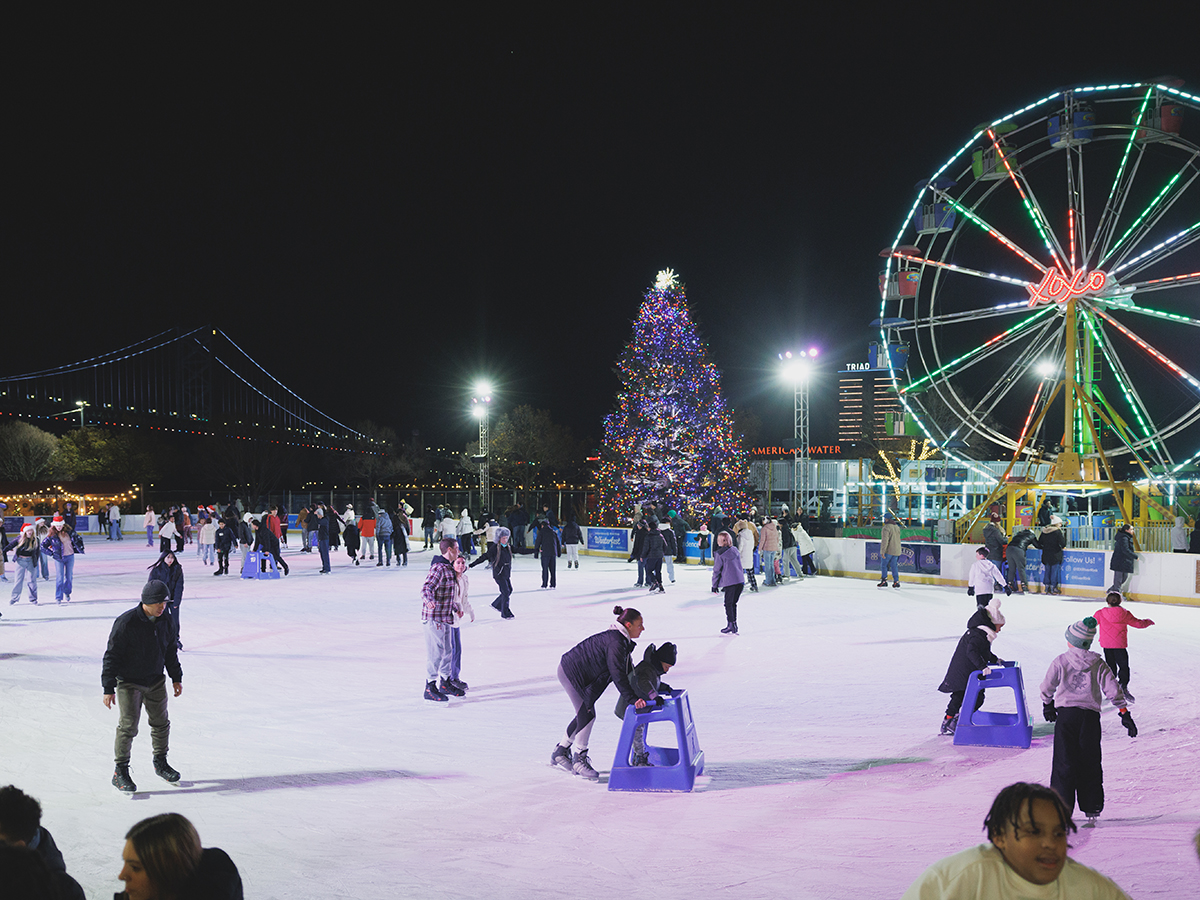 People ice skate on a white skating rink with colorful lights and a lit-up Ferris wheel in the background
