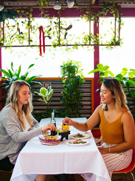 Two women share chips and tacos at a plant-filled outdoor dining space, with other guests sitting at tables nearby.