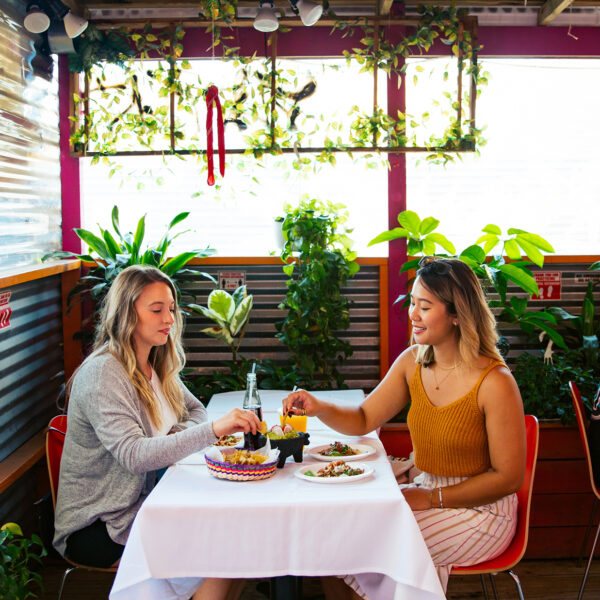 Two women share chips and tacos at a plant-filled outdoor dining space, with other guests sitting at tables nearby.