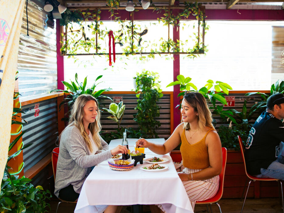 Two women share chips and tacos at a plant-filled outdoor dining space, with other guests sitting at tables nearby.