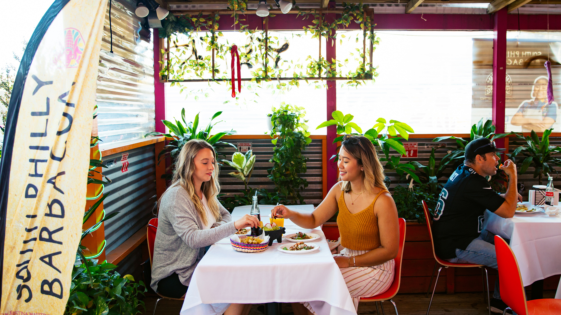 Dos mujeres comparten patatas fritas y tacos en un comedor al aire libre lleno de plantas, con otros comensales sentados en mesas cercanas.