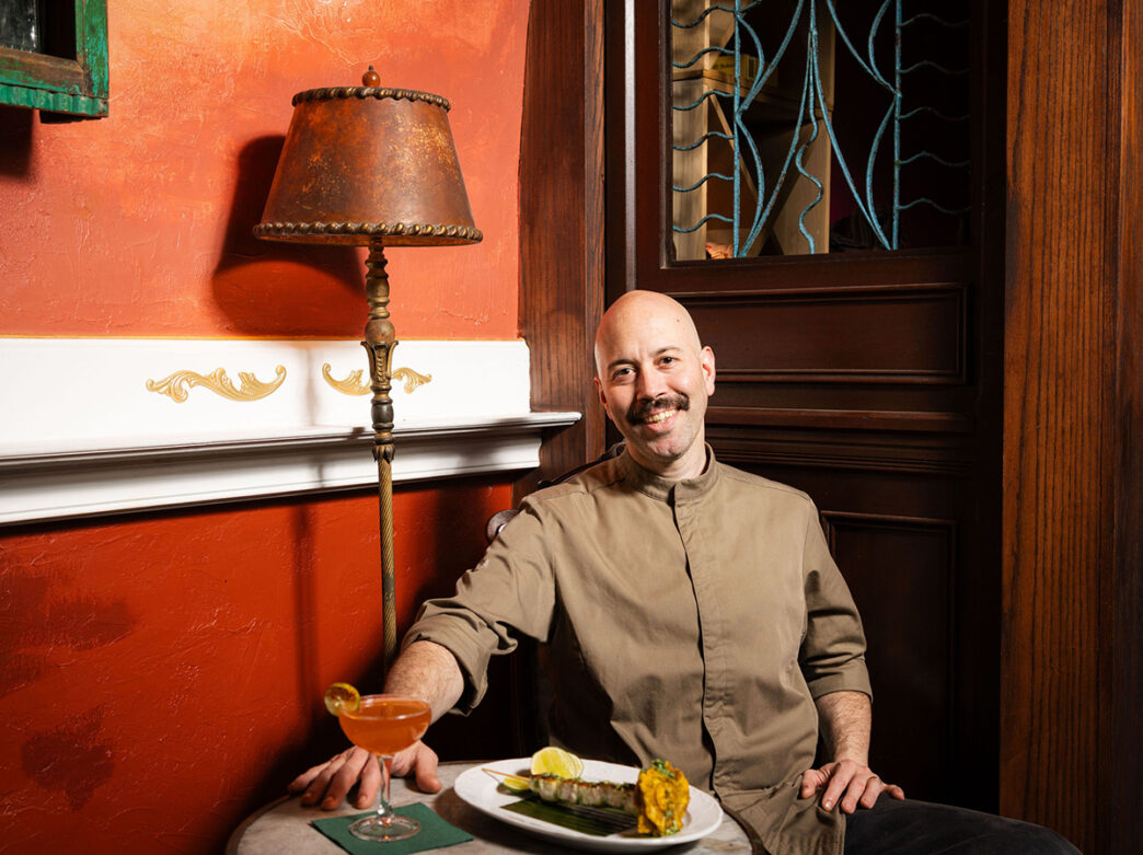 Un cocinero con calva y bigote se sienta en una mesita con un cóctel y un plato emplatado delante.
