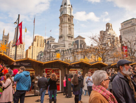 Crowds browse festive wooden stalls decorated with lights and star-shaped lanterns, with City Hall rising in the background.