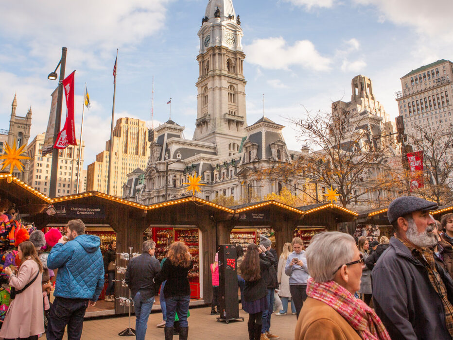 Crowds browse festive wooden stalls decorated with lights and star-shaped lanterns, with City Hall rising in the background.