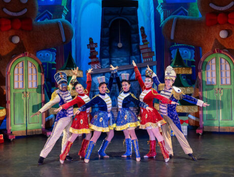 Performers dressed as festive toy soldiers in red, blue and gold costumes stand in a V formation with arms outstretched on a brightly colored holiday-themed stage featuring oversized gingerbread figures and Christmas trees.