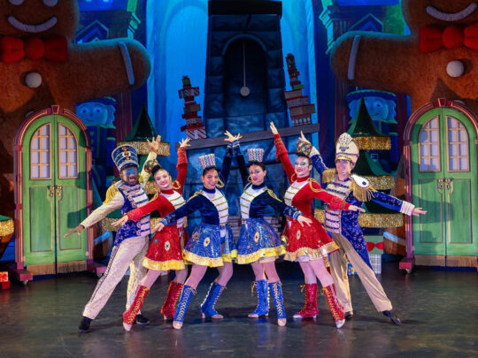 Performers dressed as festive toy soldiers in red, blue and gold costumes stand in a V formation with arms outstretched on a brightly colored holiday-themed stage featuring oversized gingerbread figures and Christmas trees.