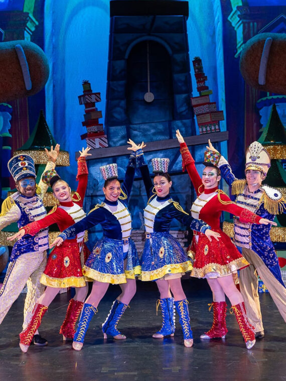 Performers dressed as festive toy soldiers in red, blue and gold costumes stand in a V formation with arms outstretched on a brightly colored holiday-themed stage featuring oversized gingerbread figures and Christmas trees.