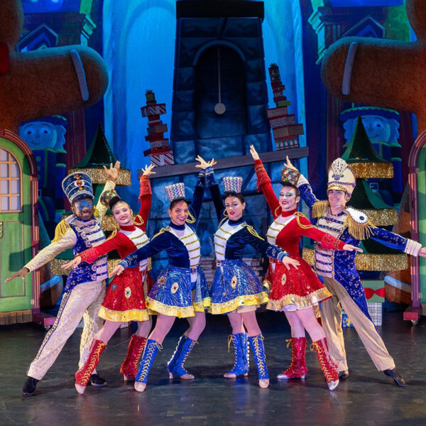 Performers dressed as festive toy soldiers in red, blue and gold costumes stand in a V formation with arms outstretched on a brightly colored holiday-themed stage featuring oversized gingerbread figures and Christmas trees.