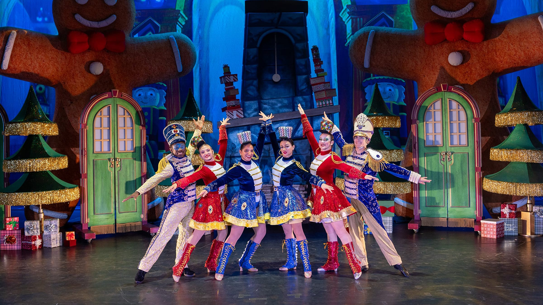 Performers dressed as festive toy soldiers in red, blue and gold costumes stand in a V formation with arms outstretched on a brightly colored holiday-themed stage featuring oversized gingerbread figures and Christmas trees.