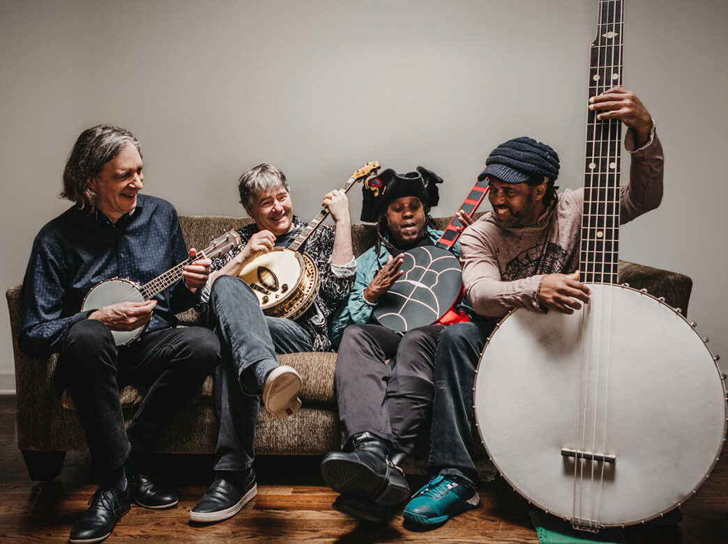 Four musicians sit on a brown couch laughing and playing various banjo-style instruments, including an oversized upright banjo.