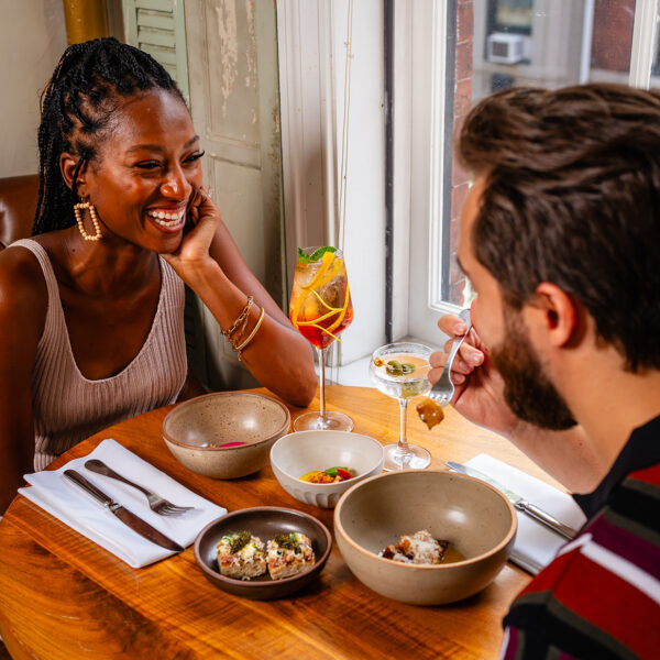 A smiling couple enjoys cocktails and small plates while sitting at a round wooden table by a sunlit window at Friday Saturday Sunday.