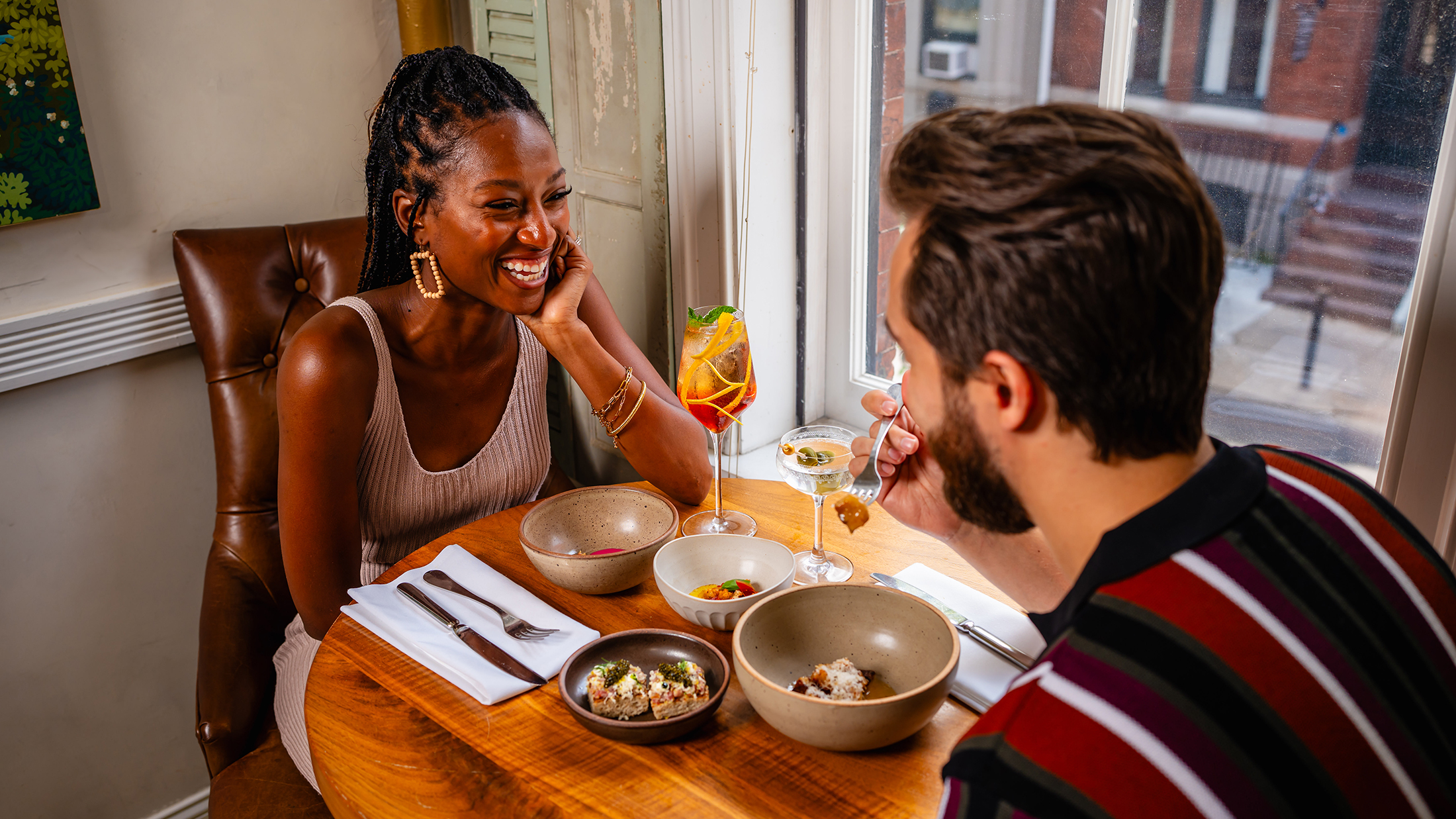 A smiling couple enjoys cocktails and small plates while sitting at a round wooden table by a sunlit window at Friday Saturday Sunday.