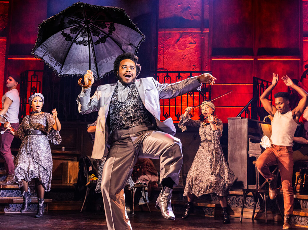 A performer in a silver suit leaps mid-dance while holding an ornate black umbrella overhead, surrounded by fellow cast members in vintage-inspirated costumes on stage.