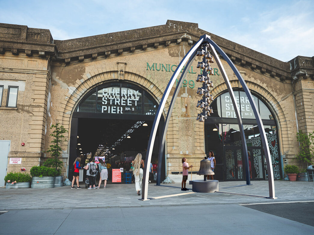 An artist's rendering of a bell tower sculpture installation in front of a warehouse-like gallery on a pier. A crowd of people walk below