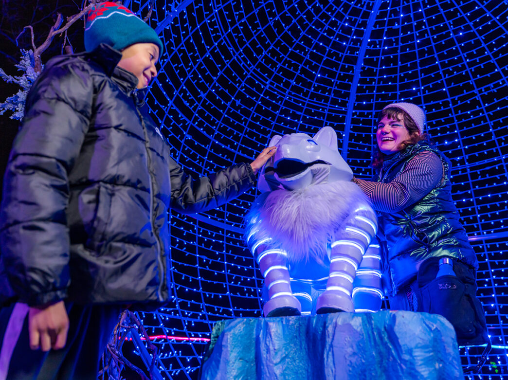 A child and an adult smile as they tough a glowing white wolf sculpture surrounded by a dome of blue lights at LumiNature.
