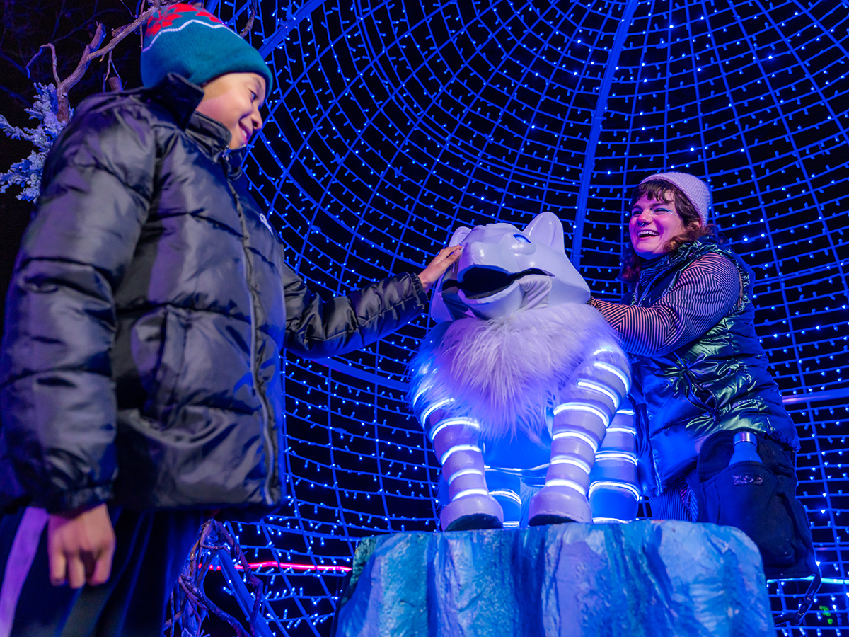 A child and an adult smile as they tough a glowing white wolf sculpture surrounded by a dome of blue lights at LumiNature.