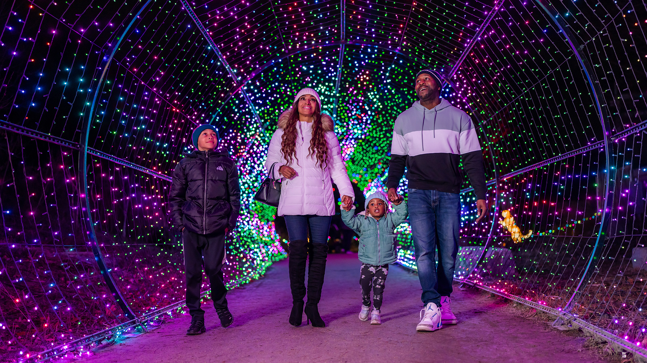 A family wearing winter jackets and beanies walks hand-in-hand through a tunnel of colorful holiday lights at night.