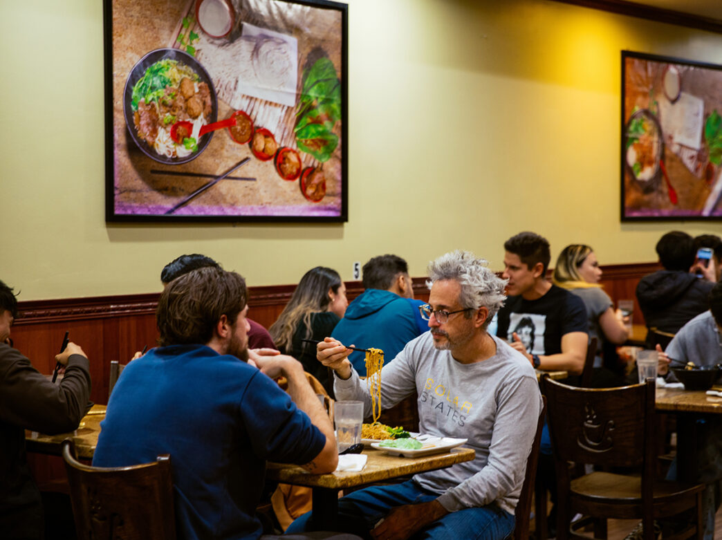 Les clients dégustent des assiettes de nouilles à l'intérieur d'un restaurant, assis à des tables en bois sous de grandes photographies culinaires accrochées aux murs.