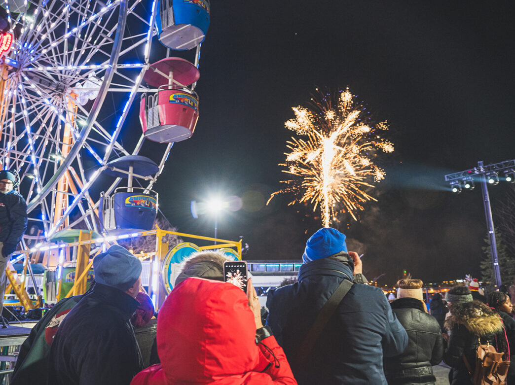 Visitors bundles in winter coasts watch fireworks burst overhead besides a glowing Ferris wheel.