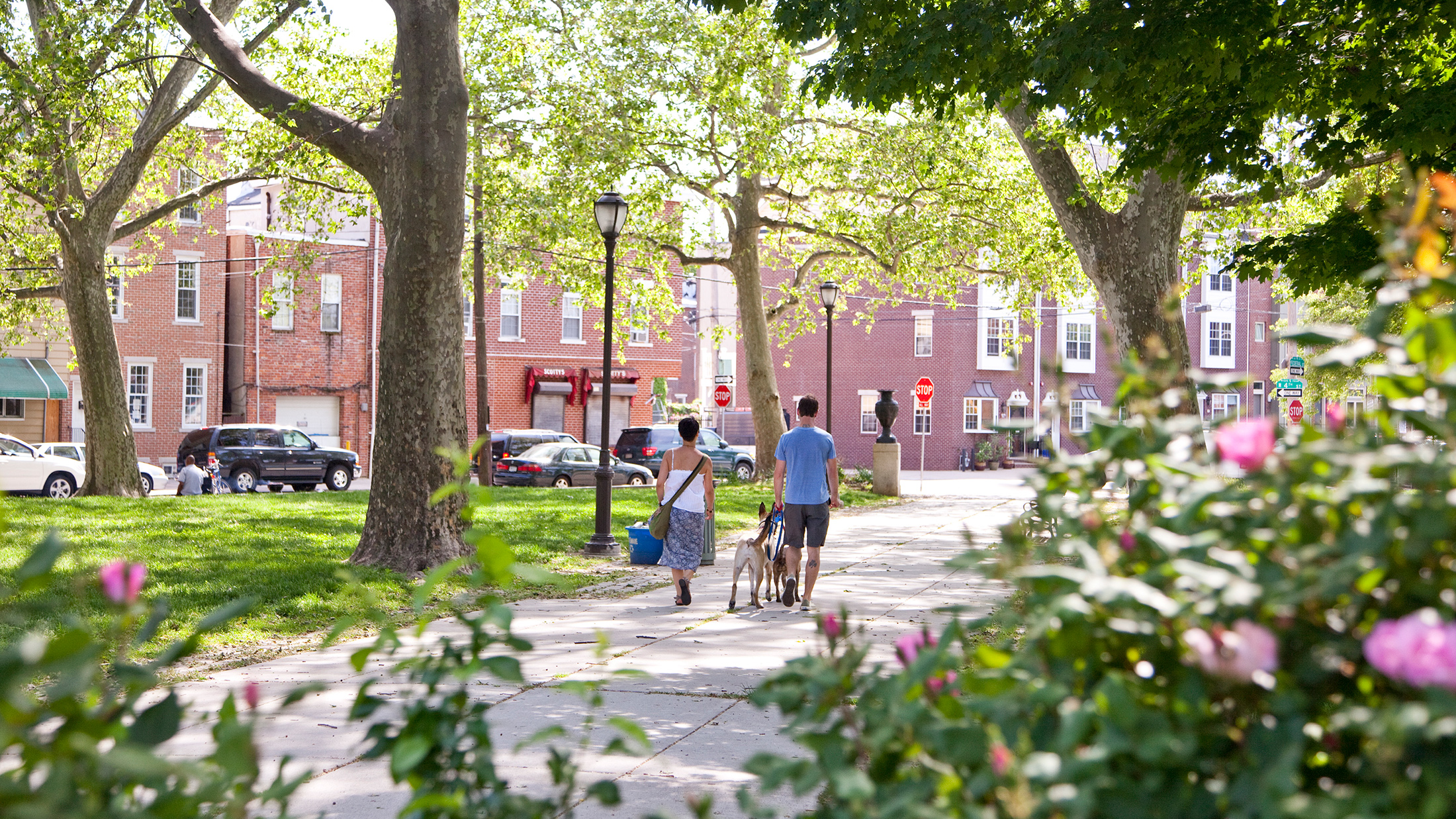 A couple walks their dogs along a sunny, tree-lined path through a park in Philadelphia's Pennsport neighborhood. Brick row homes and cars parked on the road are in the background.