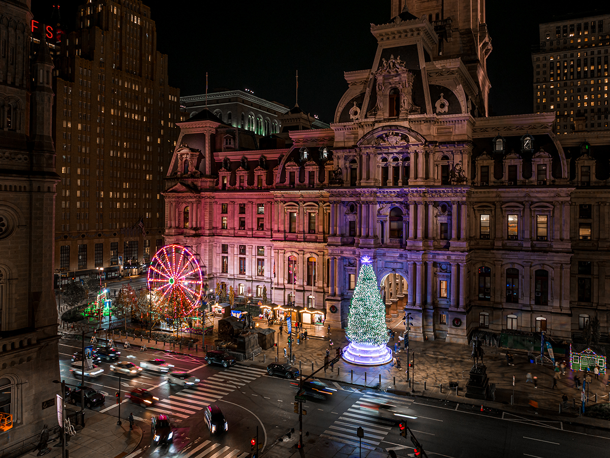 An aerial nighttime view of Philadelphia City Hall glowing with colorful up lighting, with a bright Ferris wheel and a towering illuminated Christmas tree decorating the plaza.