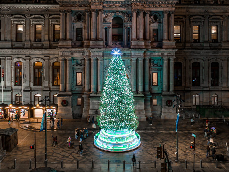 A large Christmas tree in front of City Hall illuminated with bright white and blue lights shines brightly against the facade of City Hall.