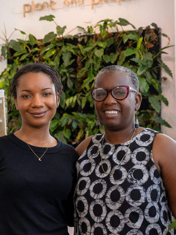 Les deux propriétaires souriants de Plant & People se tiennent à l'intérieur de leur boutique lumineuse et remplie de plantes, entourés de verdure, de cadeaux et de la lumière naturelle qui pénètre à flots par les vitrines.