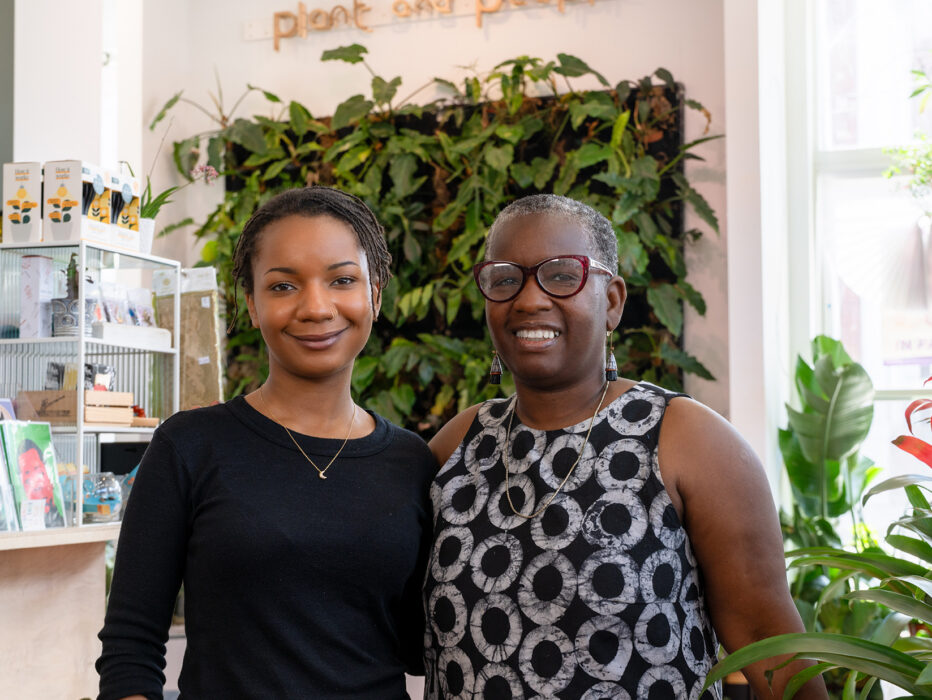 Two smiling owners of Plant & People stand inside their bright, plant-filled shop, surrounded by greenery, gifts and natural light streaming through the windows.