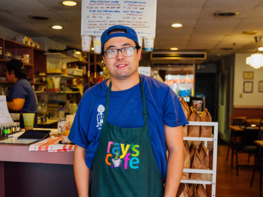 Un empleado con camisa azul y delantal negro de Ray's Cafe está de pie frente al mostrador de una acogedora cafetería, con estanterías de ingredientes y un menú escrito a mano detrás de él.