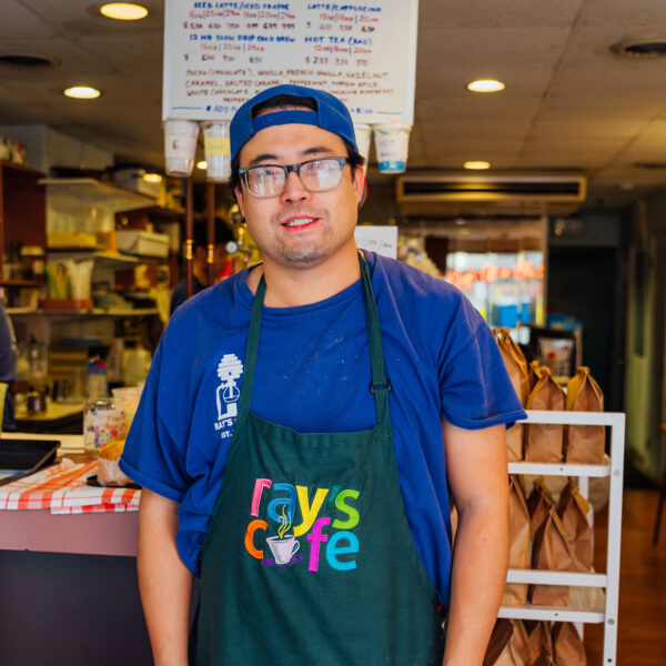 A staff member wearing a blue shirt and black Ray's Cafe apron stands in front of the counter in a cozy cafe, with shelves of ingredients and handwritten menu board behind him.
